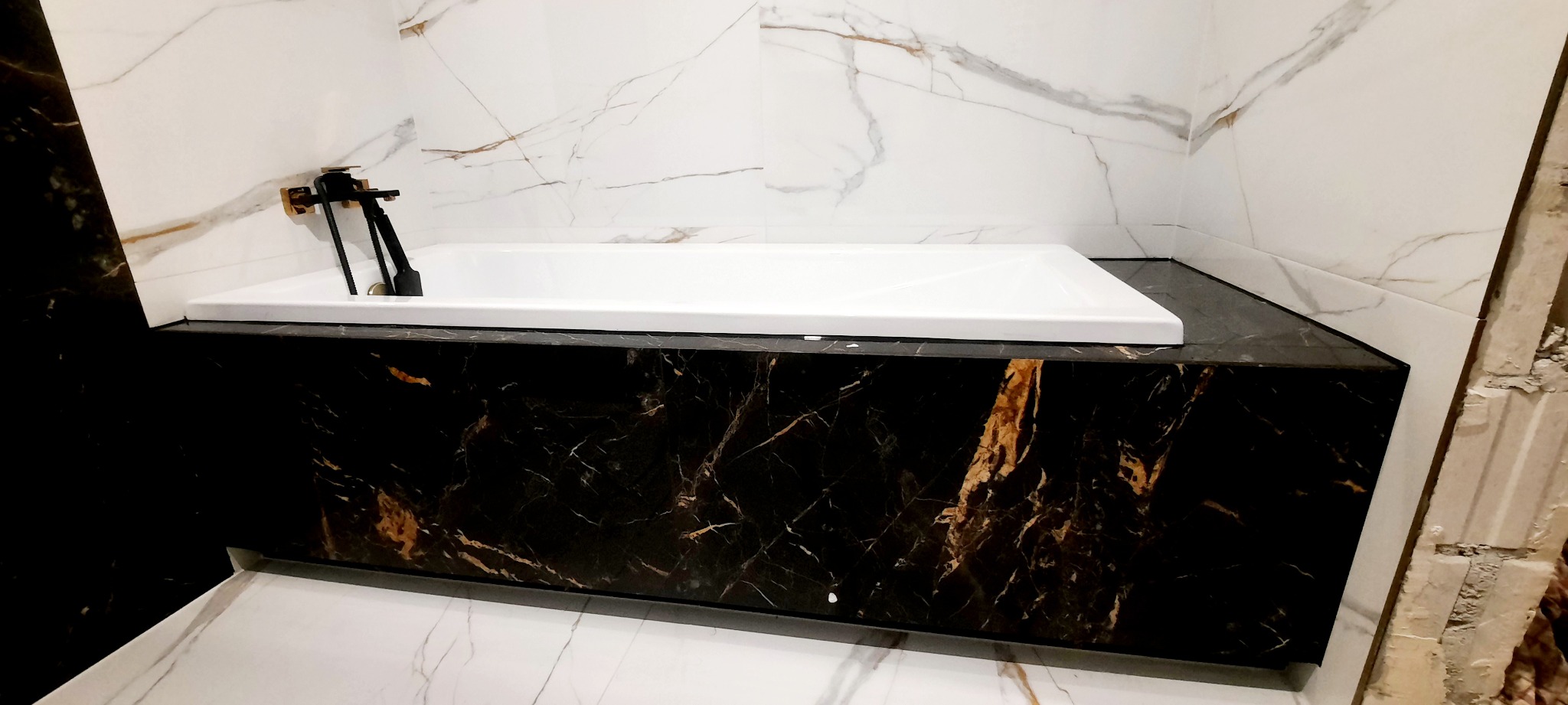 Biała wanna obudowana czarnym marmurem z złotymi żyłkami, czarna bateria, białe marmurowe płytki na ścianach i podłodze, fragment ściany w trakcie wykończenia.