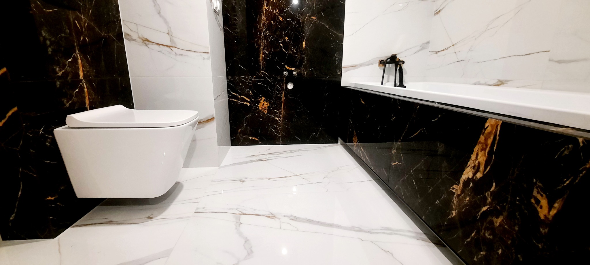 Nowoczesna łazienka z białymi marmurkowymi płytkami podłogowymi i ścianami częściowo wyłożonymi czarnym marmurem z złotymi żyłami, wisząca toaleta i zabudowana wanna z czarną armaturą.