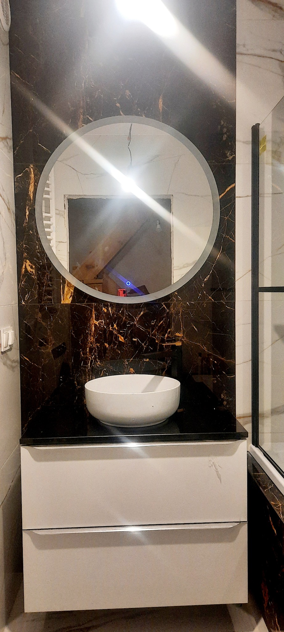 Umywalka nablatowa, okrągłe lustro z podświetleniem i marmurowe płytki w ciemnych barwach z złotymi żyłami, kabina prysznicowa w tle.