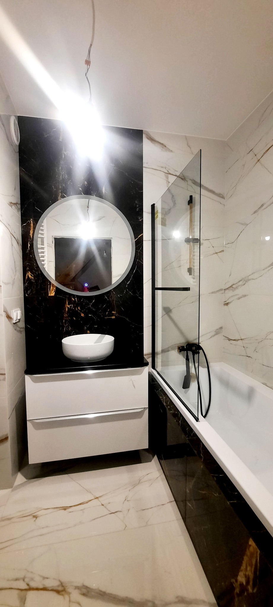 Nowoczesna łazienka z białymi i czarnymi marmurowymi płytkami, okrągłym lustrem z podświetleniem LED nad białą szafką z umywalką i czarną armaturą przy wannie z parawanem.
