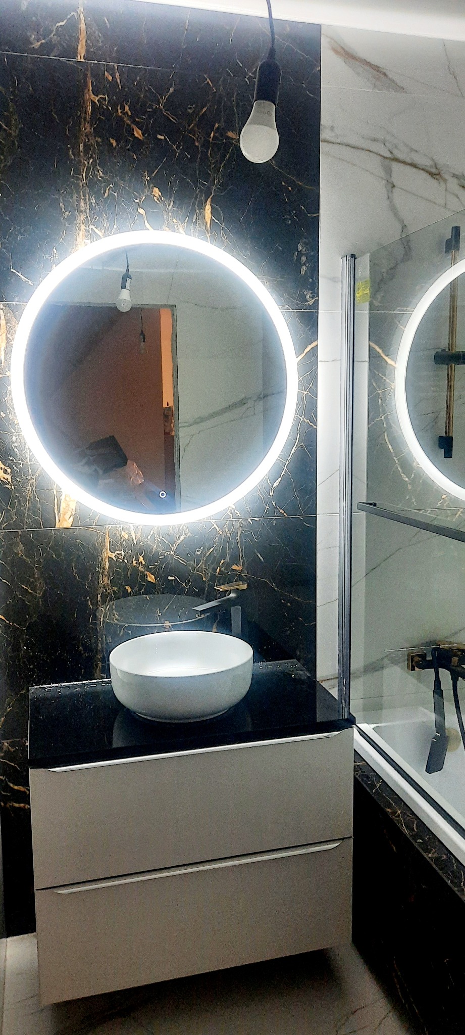 Nowoczesna łazienka z czarnym marmurem, okrągłym lustrem z podświetleniem LED, białą umywalką nablatową i szarą szafką pod umywalką. W odbiciu lustra widoczny fragment innego pomieszczenia.