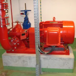 Czerwona pompa przemysłowa z silnikiem elektrycznym na betonowym fundamencie, widoczne rury i zawory w kolorach czerwonym i niebieskim, metalowa listwa perforowana w tle.