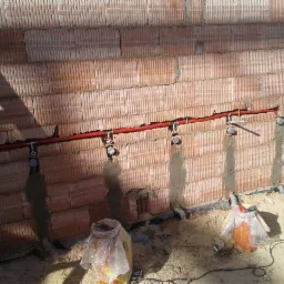 Czerwona rura instalacyjna zamocowana do ceglanej ściany z widocznymi uchwytami i fragmentami instalacji, w tle piasek i narzędzia pracy.