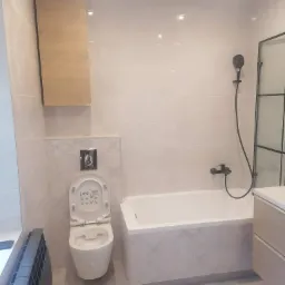 Jasna łazienka po renowacji z wiszącą toaletą z otwartą klapą, zabudowaną wanną z czarną armaturą, grzejnikiem żeberkowym i szafką wiszącą.