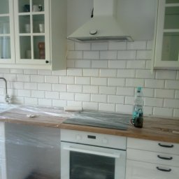 WAX-BUD Usługi remontowo-budowlane Maciej Wawer - Biała kuchnia w trakcie zabezpieczania folią malarską przed pracami wykończeniowymi, widoczne białe szafki z przeszklonymi frontami, okap, białe płytki na ścianie, blat drewniany, butelka wody...