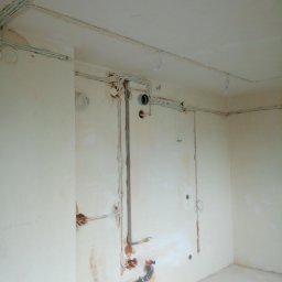 WAX-BUD Usługi remontowo-budowlane Maciej Wawer - Wnętrze w trakcie remontu: widoczne instalacje elektryczne i hydrauliczne, ściany przygotowane pod gładzie, rury kanalizacyjne przy podłodze.