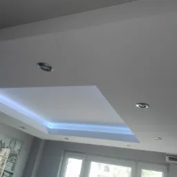 Podwieszany sufit z oświetleniem LED w kolorze chłodnego błękitu, widoczne okrągłe halogeny, ściana z obrazem w tle.