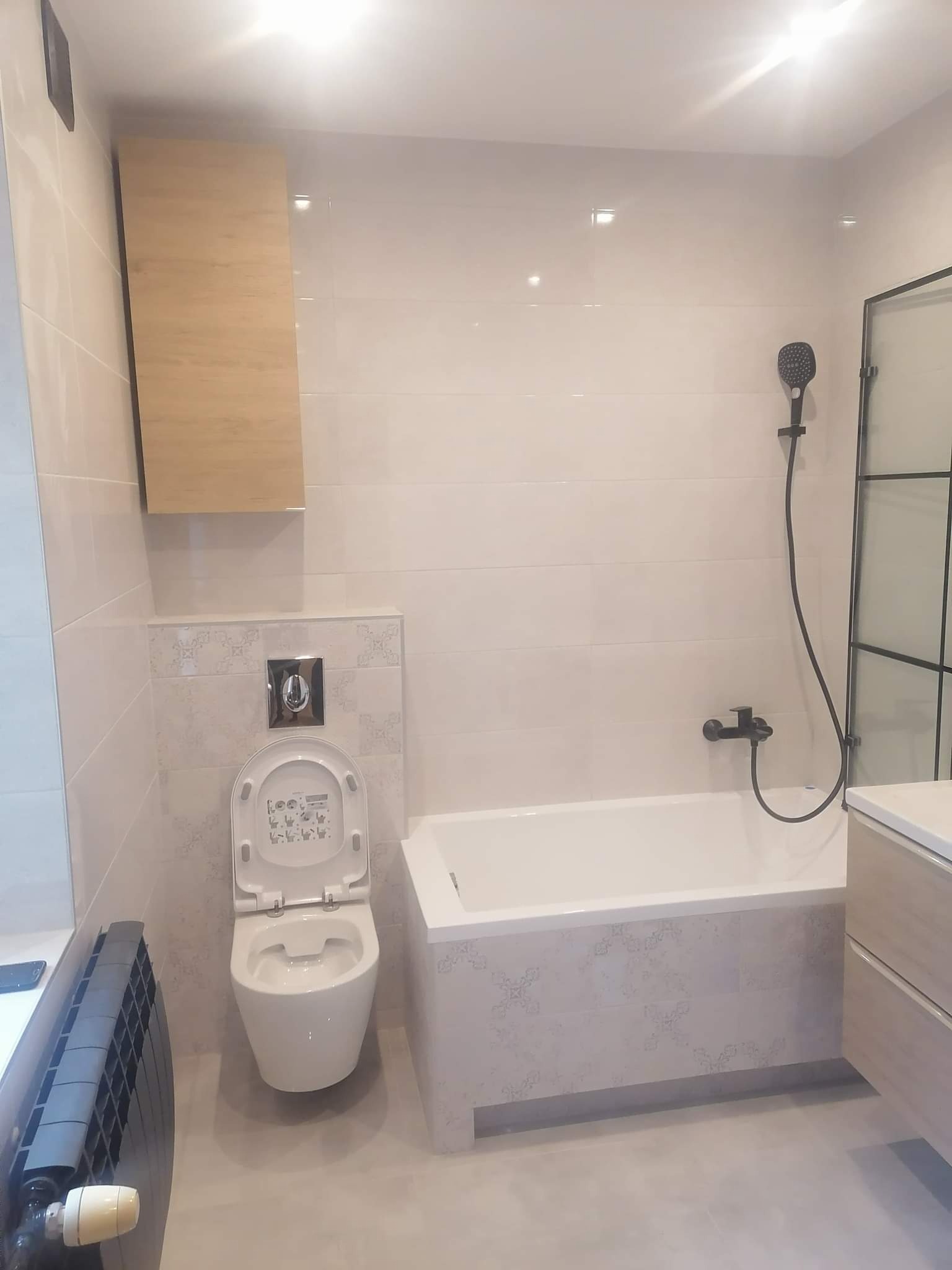 Jasna łazienka po renowacji z wiszącą toaletą z otwartą klapą, zabudowaną wanną z czarną armaturą, grzejnikiem żeberkowym i szafką wiszącą.