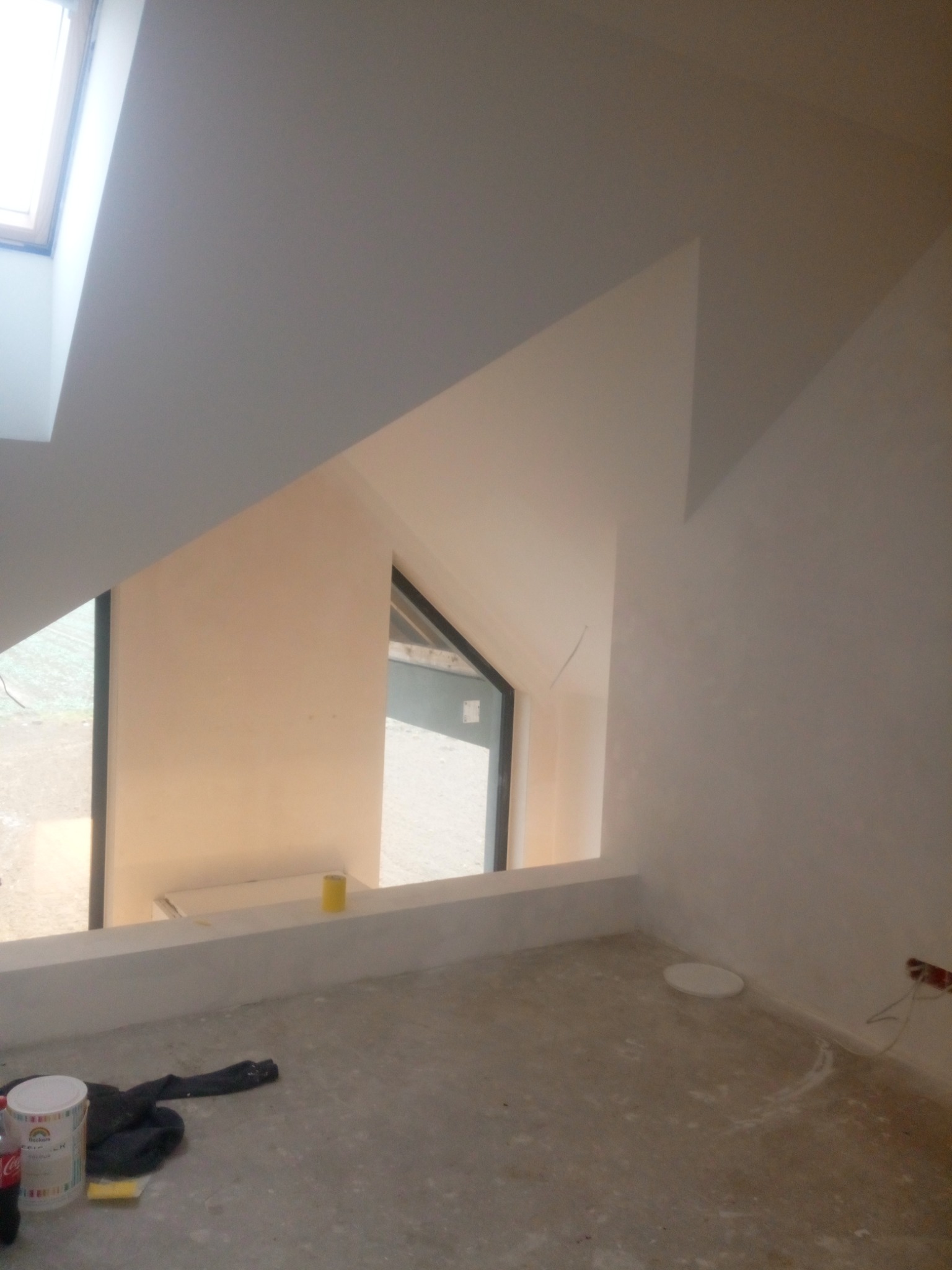 Pokój na poddaszu w trakcie wykańczania, widoczne okno dachowe, ściany przygotowane pod malowanie, na podłodze wiadro z farbą i folia ochronna.
