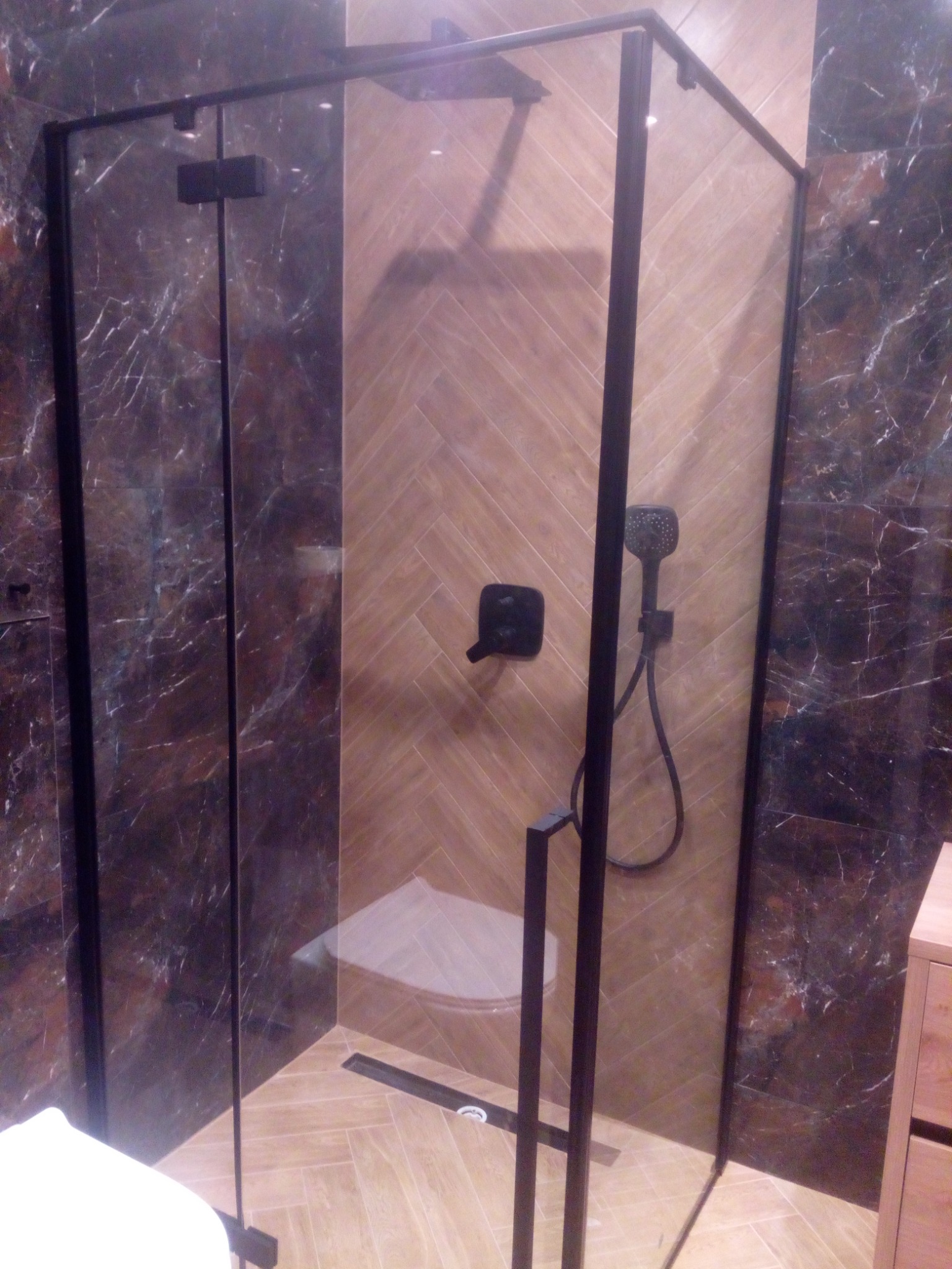 Nowoczesna kabina prysznicowa z czarnymi profilami i armaturą na tle ściany z płytek ułożonych w jodełkę i marmurowych płytek.