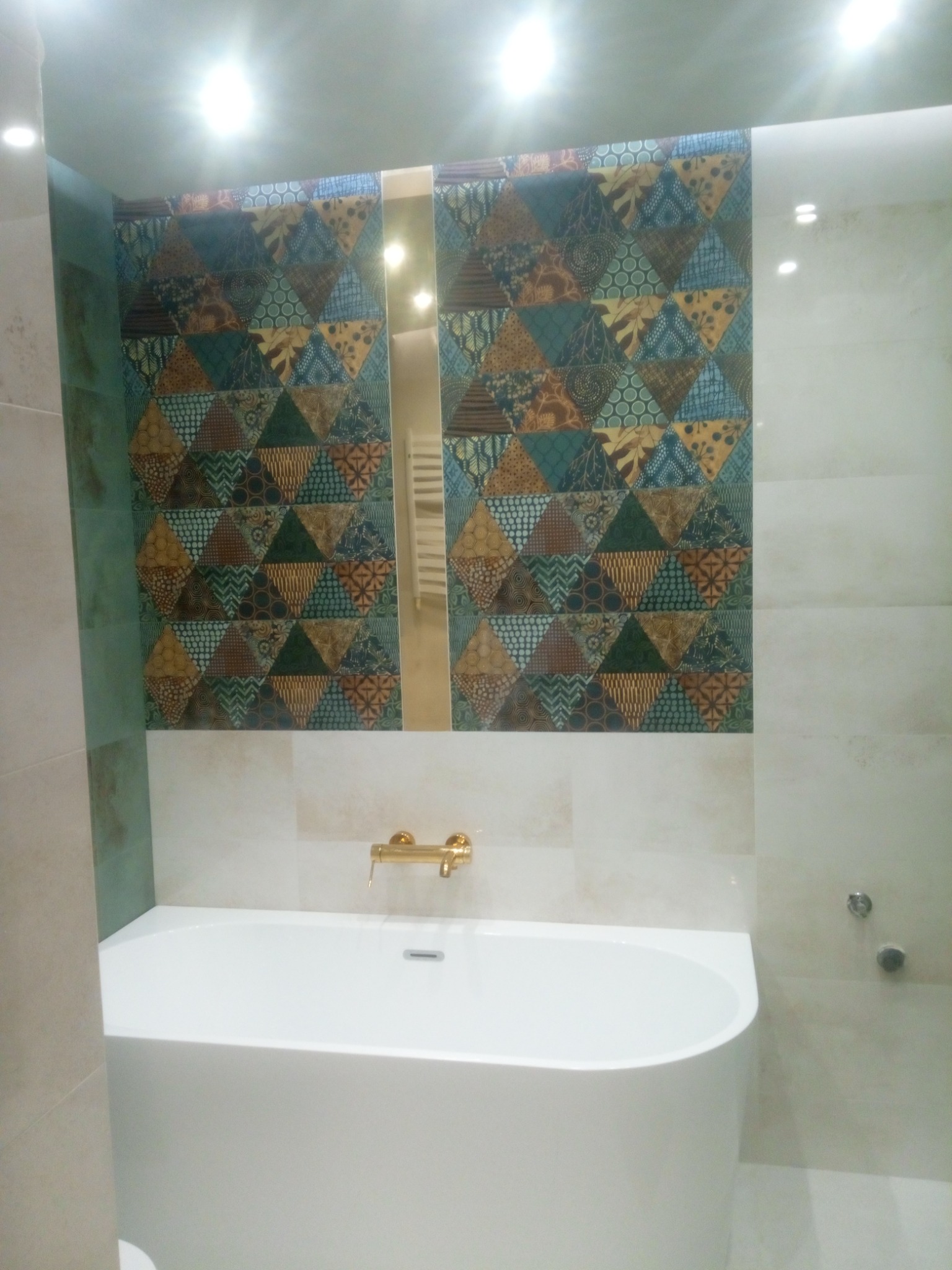 Nowoczesna łazienka z białą wanną, złotą baterią i dekoracyjną ścianą z płytek w geometryczne wzory, odbijająca się w lustrze.