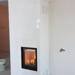 Biały, kaflowy kominek z paleniskiem i dekoracyjną figurką kota na górze, widoczny w surowym wnętrzu z szarą podłogą i śladami po instalacji na ścianie.