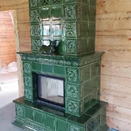 Nowoczesny, zielony piec kaflowy z dekoracyjnymi wzorami, zwieńczony figurkami kotów, w surowym wnętrzu drewnianego domu w trakcie budowy.