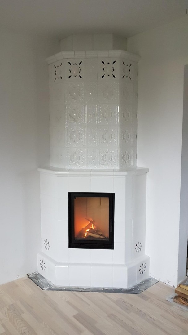 Biały, kaflowy piec kominkowy narożny z paleniskiem, widoczne płomienie, dekoracyjne płytki z wzorami, w tle białe ściany i jasna podłoga.