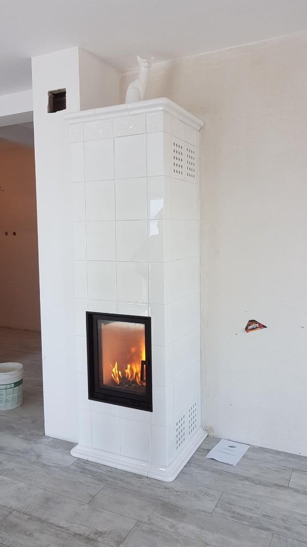 Biały, kaflowy kominek z paleniskiem i dekoracyjną figurką kota na górze, widoczny w surowym wnętrzu z szarą podłogą i śladami po instalacji na ścianie.