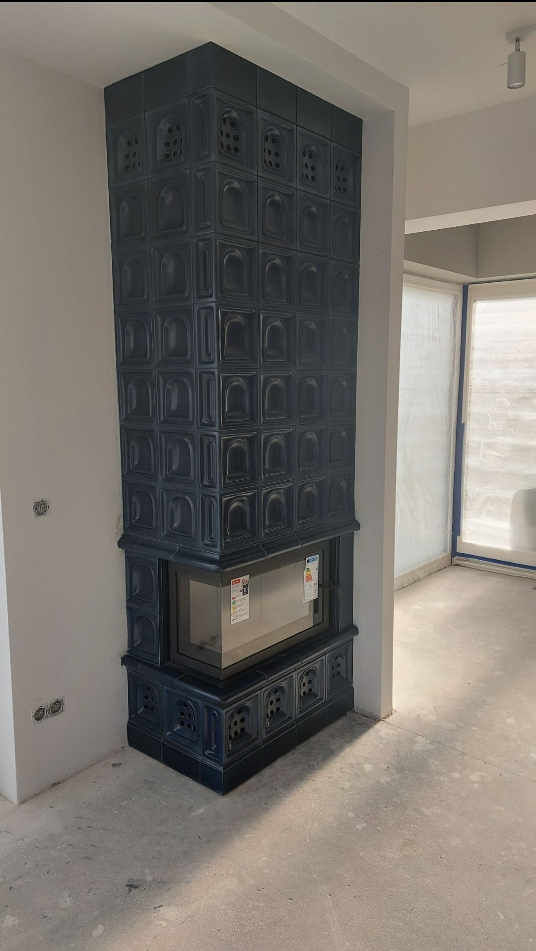 Narożny, niebieski piec kaflowy z szybą, w surowym wnętrzu z widocznymi gniazdkami elektrycznymi i oknem zabezpieczonym folią.