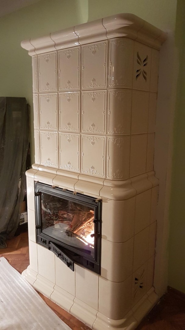 Beżowy piec kaflowy z otwartymi drzwiczkami paleniska, w którym widać płomienie ognia, dekoracyjne płytki z motywem roślinnym i wywietrznik w kształcie stylizowanej rozety.