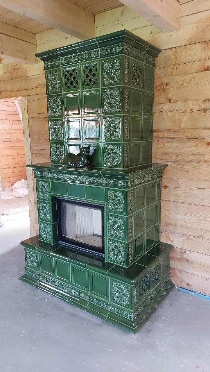 Nowoczesny, zielony piec kaflowy z dekoracyjnymi wzorami, zwieńczony figurkami kotów, w surowym wnętrzu drewnianego domu w trakcie budowy.