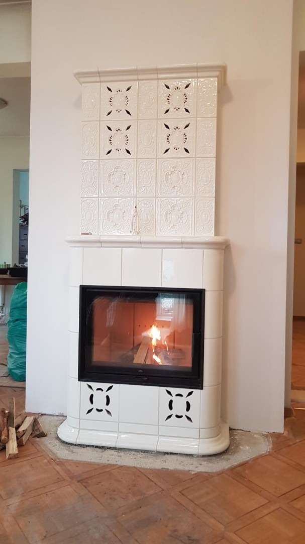 Biały, kaflowy piec kominkowy z palącym się drewnem, dekoracyjne płytki z motywem roślinnym, drewniana podłoga w trakcie wykańczania.