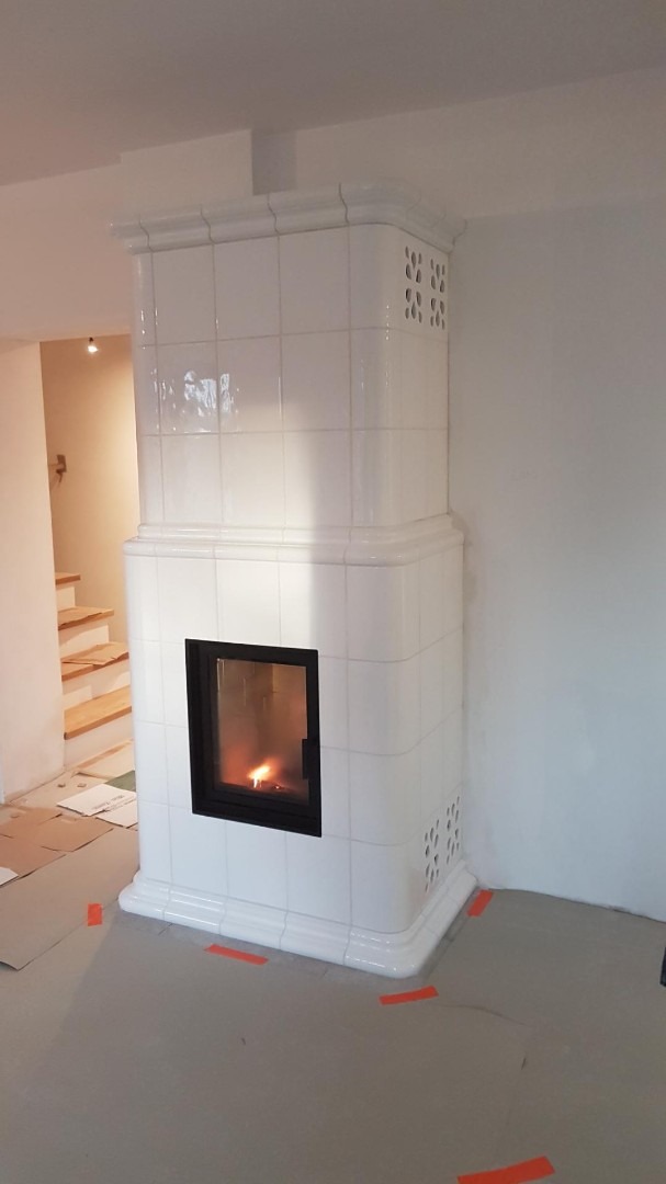 Biały, kaflowy piec kominkowy z czarnym wkładem, widoczne płomienie, wykończony ozdobnymi listwami, na podłodze zabezpieczenia budowlane.