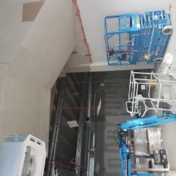 Montaż płyty karton gips na suficie w surowym wnętrzu, widoczne podnośniki koszowe i instalacje wentylacyjne.