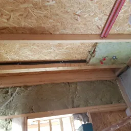 Poddasze w trakcie ocieplania wełną mineralną, widoczne drewniane belki konstrukcyjne, płyta OSB oraz instalacja elektryczna i rury grzewcze.