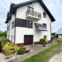 Zakład Remontowo-Budowlany Adrian Szymański - Dwupiętrowy dom jednorodzinny z balkonem, panelami słonecznymi na elewacji, wykończony w bieli z brązowymi akcentami, otoczony zielenią i kamienną ścieżką.