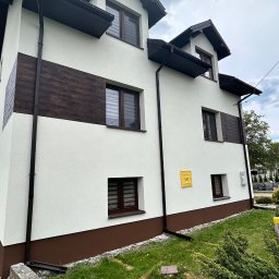 Zakład Remontowo-Budowlany Adrian Szymański - Dwupiętrowy dom z białą elewacją, brązowymi akcentami imitującymi drewno, ciemnymi oknami i rynnami, widoczna tabliczka 'GAZ', na tle pochmurnego nieba i fragmentu zieleni.