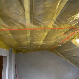 Zakład Remontowo-Budowlany Adrian Szymański - Sufit poddasza zabezpieczony folią budowlaną przed ociepleniem, widoczne drewniane belki konstrukcyjne i pomarańczowa taśma mocująca, surowe ściany w tle.