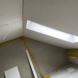 Zakład Remontowo-Budowlany Adrian Szymański - Montaż płyt kartonowo-gipsowych na poddaszu ze świetlikiem, widoczne profile, izolacja i fragment ściany z cegły. W tle widoczny surowy tynk.