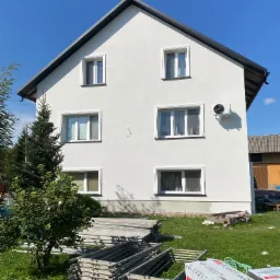 Dwupiętrowy dom z białą elewacją, widoczne okna z białymi ramami, klimatyzator na ścianie, na trawniku leżące rusztowania i paczki styropianu, błękitne niebo.