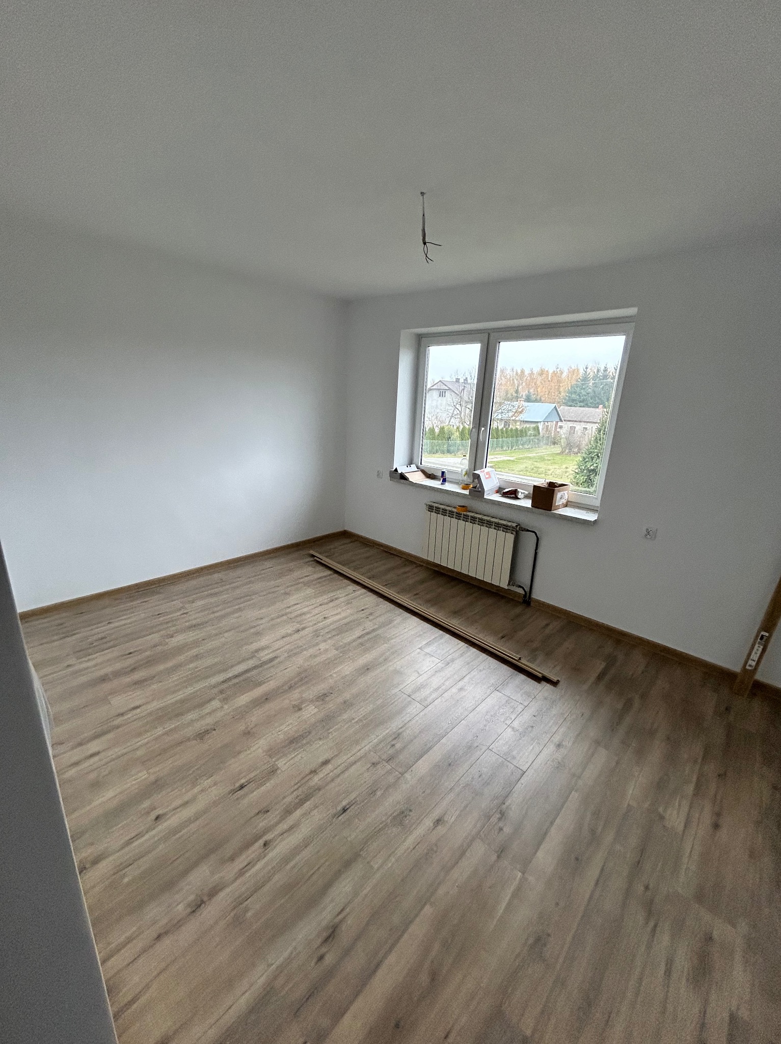 Pokój po remoncie z ułożoną podłogą z paneli, białe ściany i okno z widokiem na domy i drzewa, listwy przypodłogowe przygotowane do montażu.