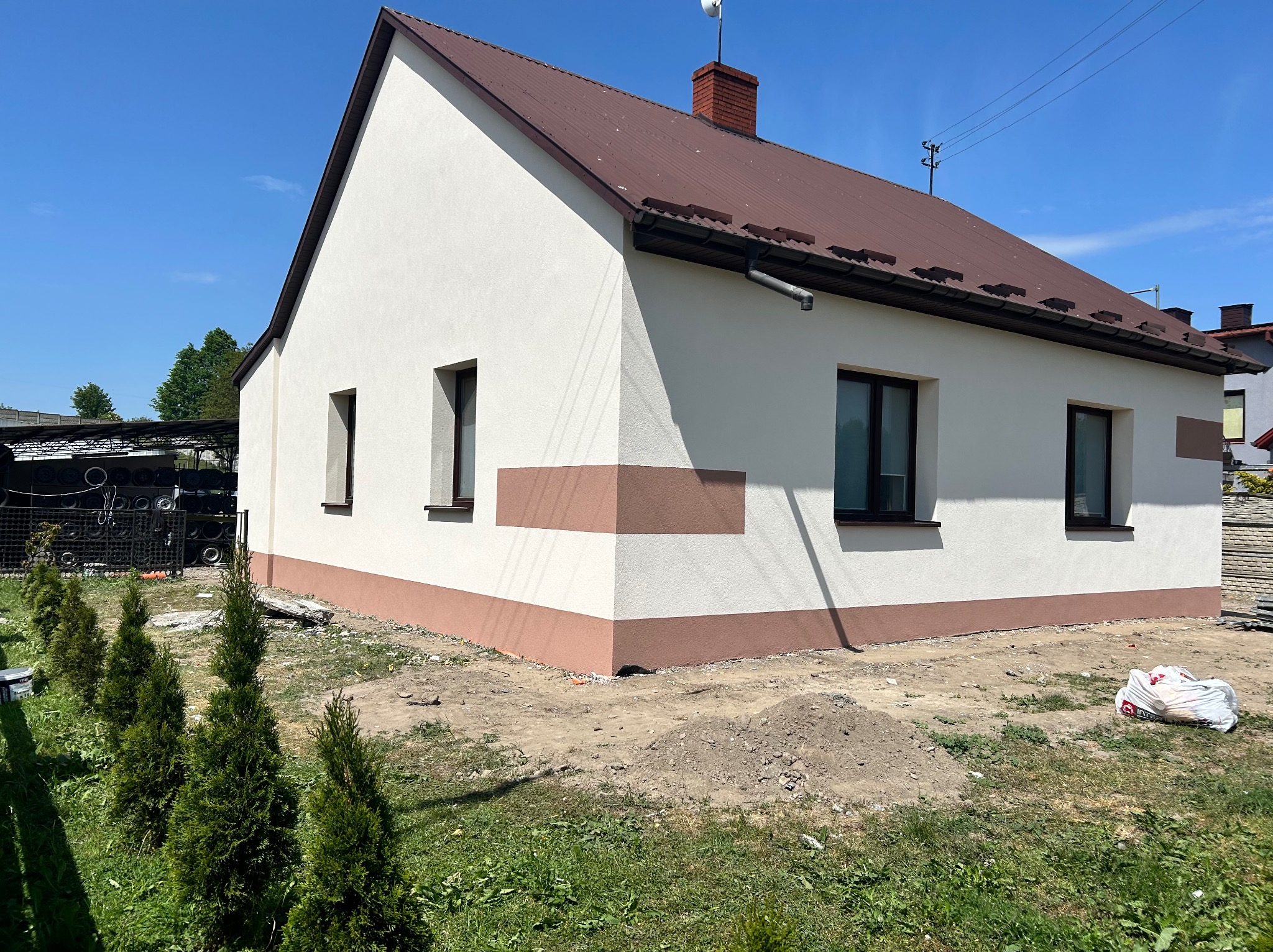 Dom jednorodzinny po tynkowaniu elewacji na biało z brązowymi akcentami, widoczne okna z ciemnymi ramami, brązowy dach i niezagospodarowany teren wokół budynku z workiem gruzu.