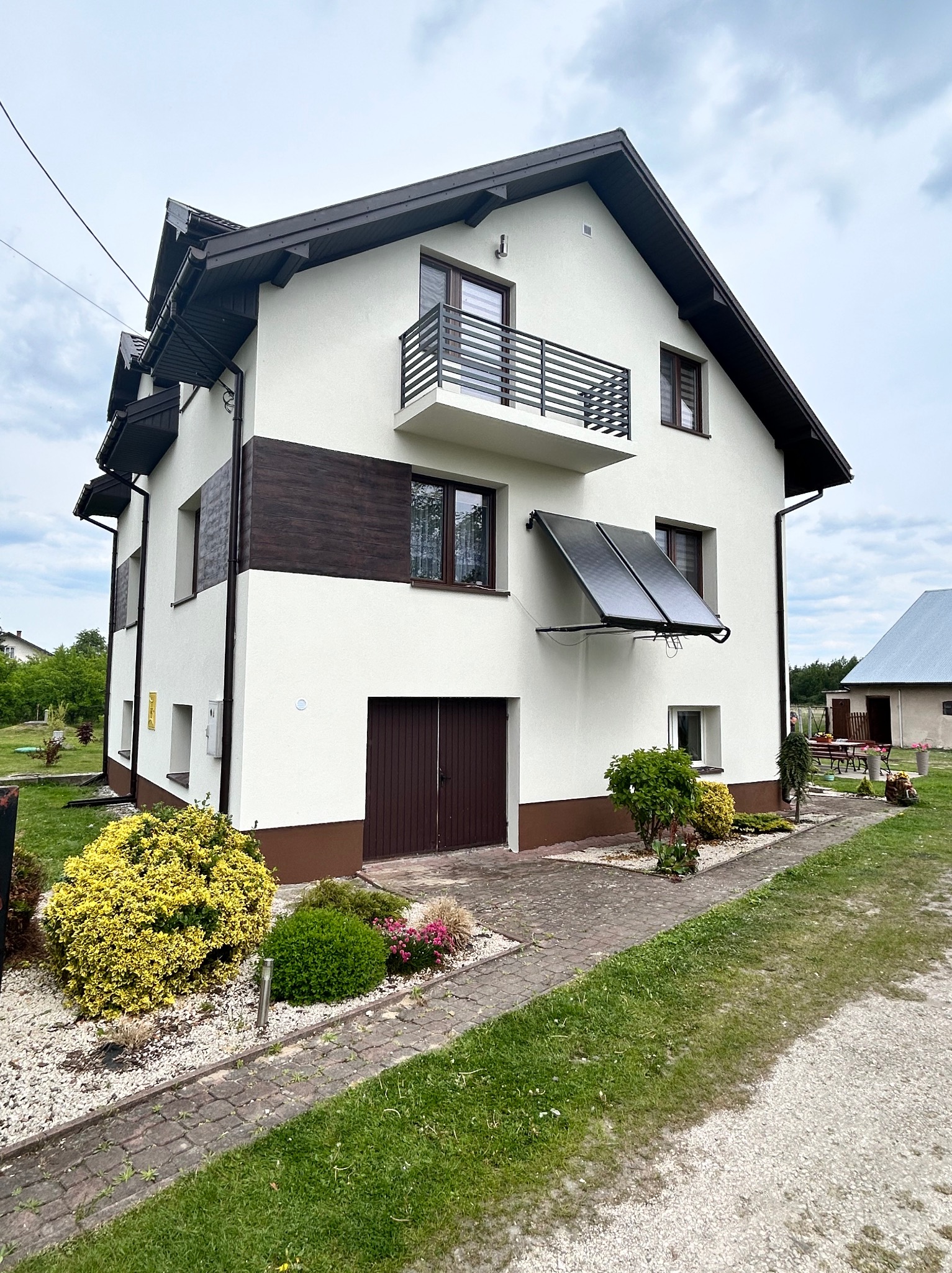 Dwupiętrowy dom jednorodzinny z balkonem, panelami słonecznymi na elewacji, wykończony w bieli z brązowymi akcentami, otoczony zielenią i kamienną ścieżką.