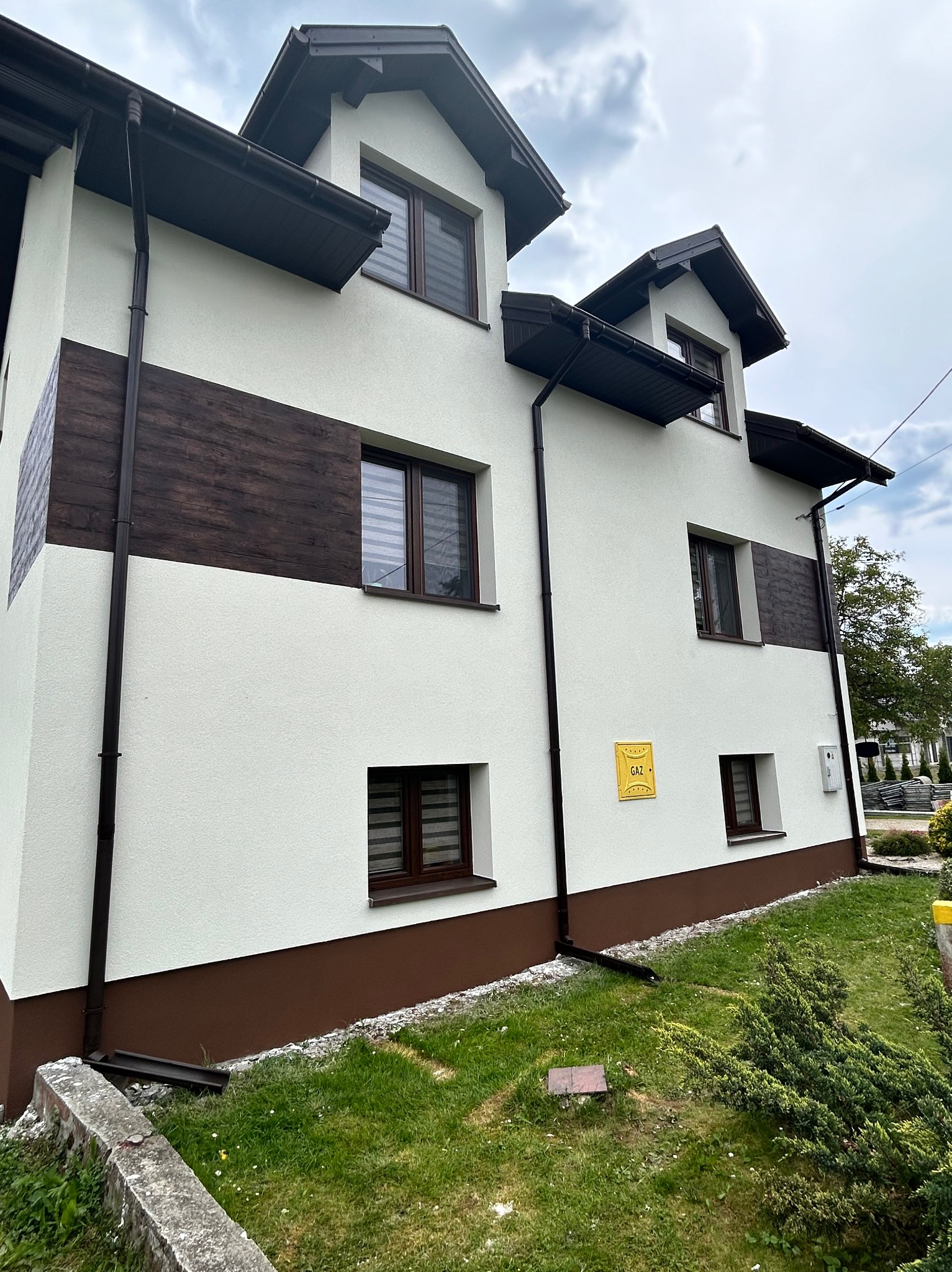 Dwupiętrowy dom z białą elewacją, brązowymi akcentami imitującymi drewno, ciemnymi oknami i rynnami, widoczna tabliczka 'GAZ', na tle pochmurnego nieba i fragmentu zieleni.