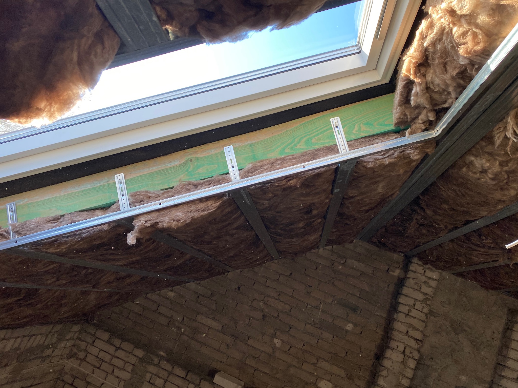 Ocieplenie poddasza wełną mineralną, widoczne krokwie, metalowe profile i okno dachowe z widokiem na niebo, ściany z cegły.