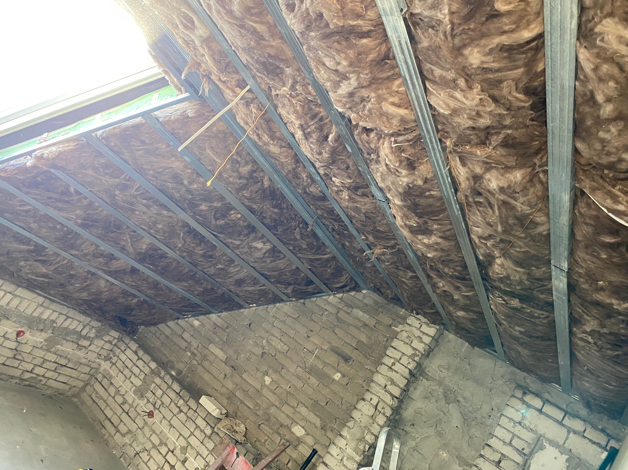 Izolacja poddasza z wełny mineralnej zamontowana na metalowym stelażu, widoczne fragmenty muru z cegły i okno dachowe.