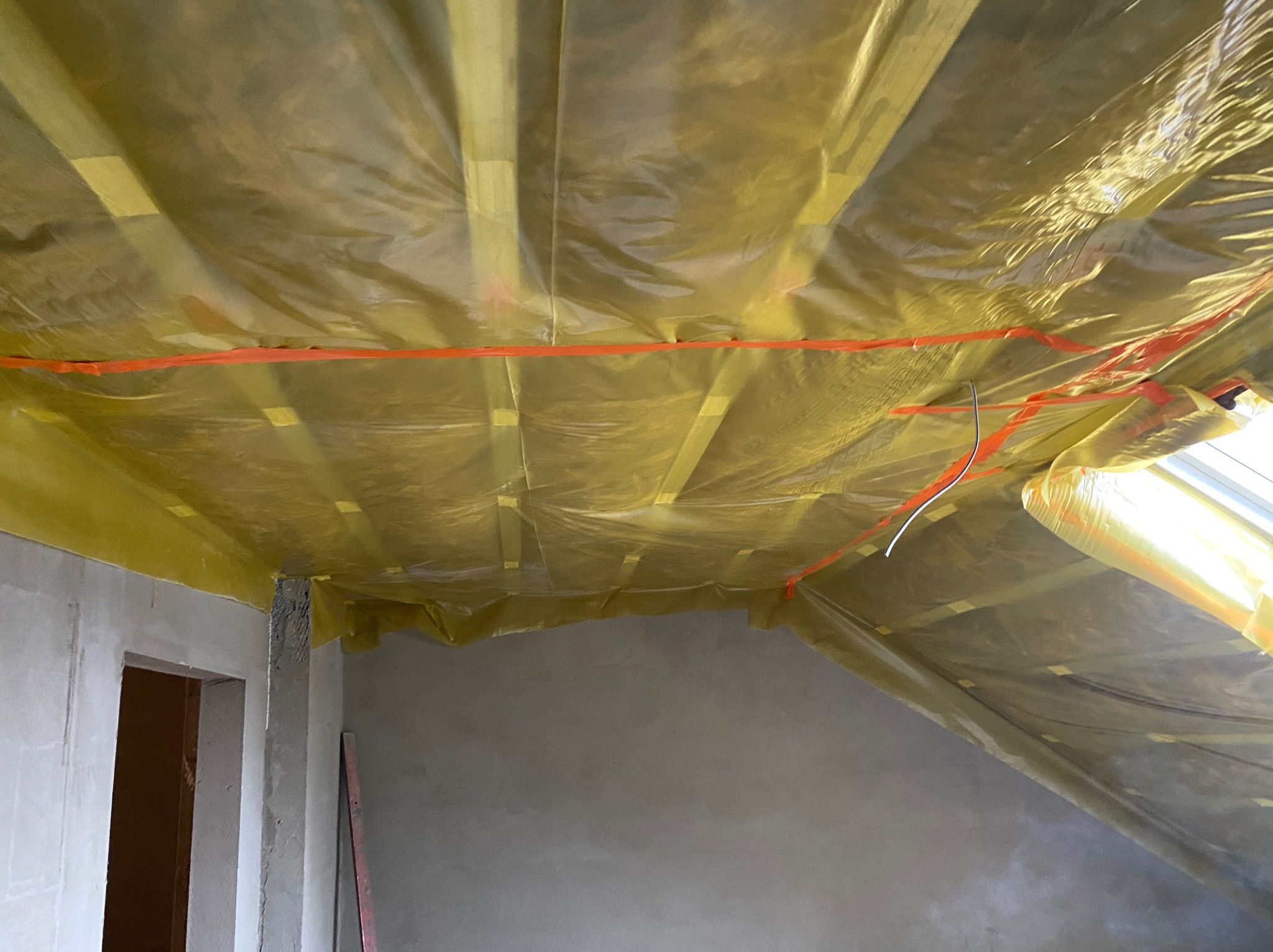 Sufit poddasza zabezpieczony folią budowlaną przed ociepleniem, widoczne drewniane belki konstrukcyjne i pomarańczowa taśma mocująca, surowe ściany w tle.
