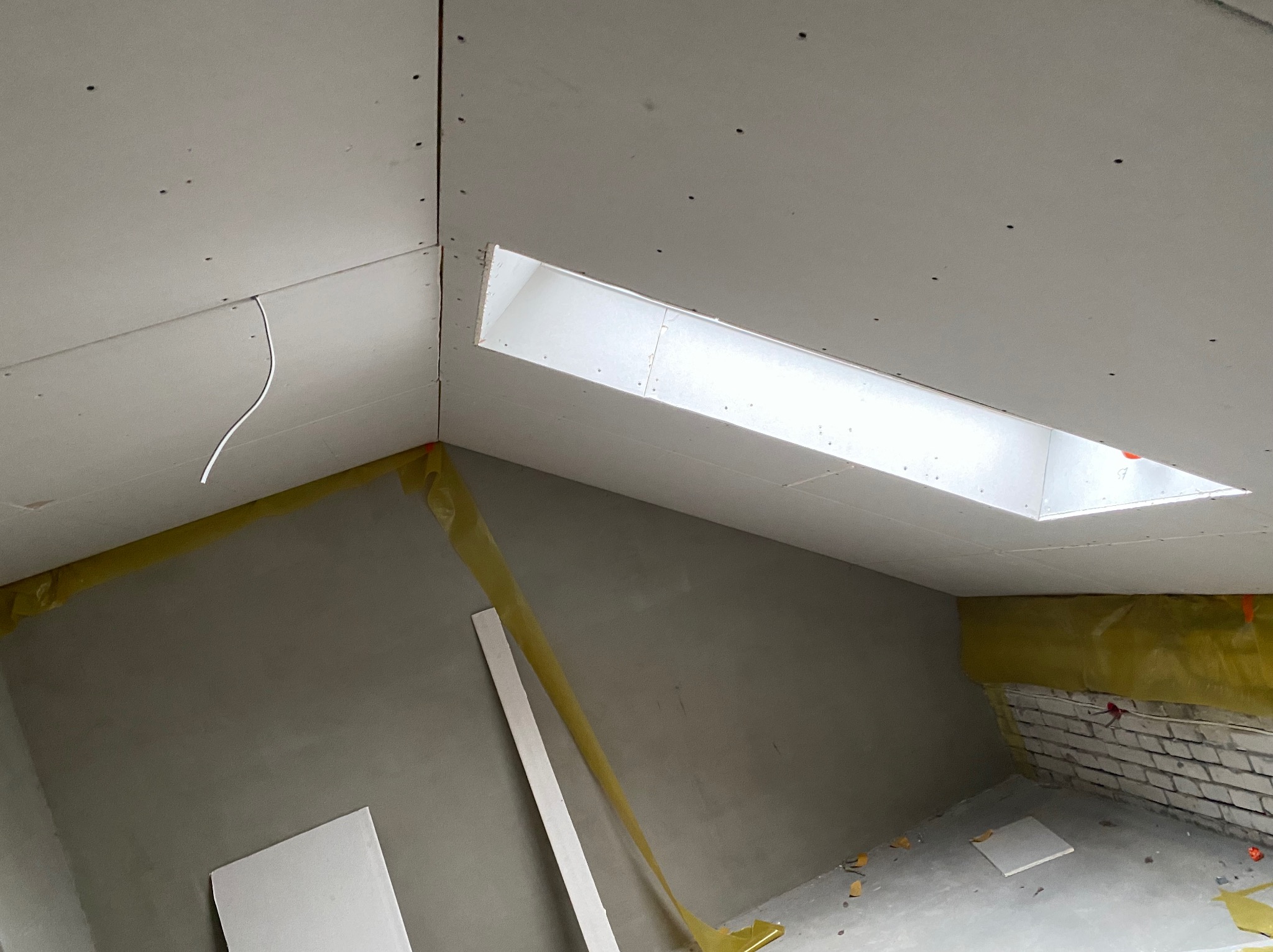 Montaż płyt kartonowo-gipsowych na poddaszu ze świetlikiem, widoczne profile, izolacja i fragment ściany z cegły. W tle widoczny surowy tynk.