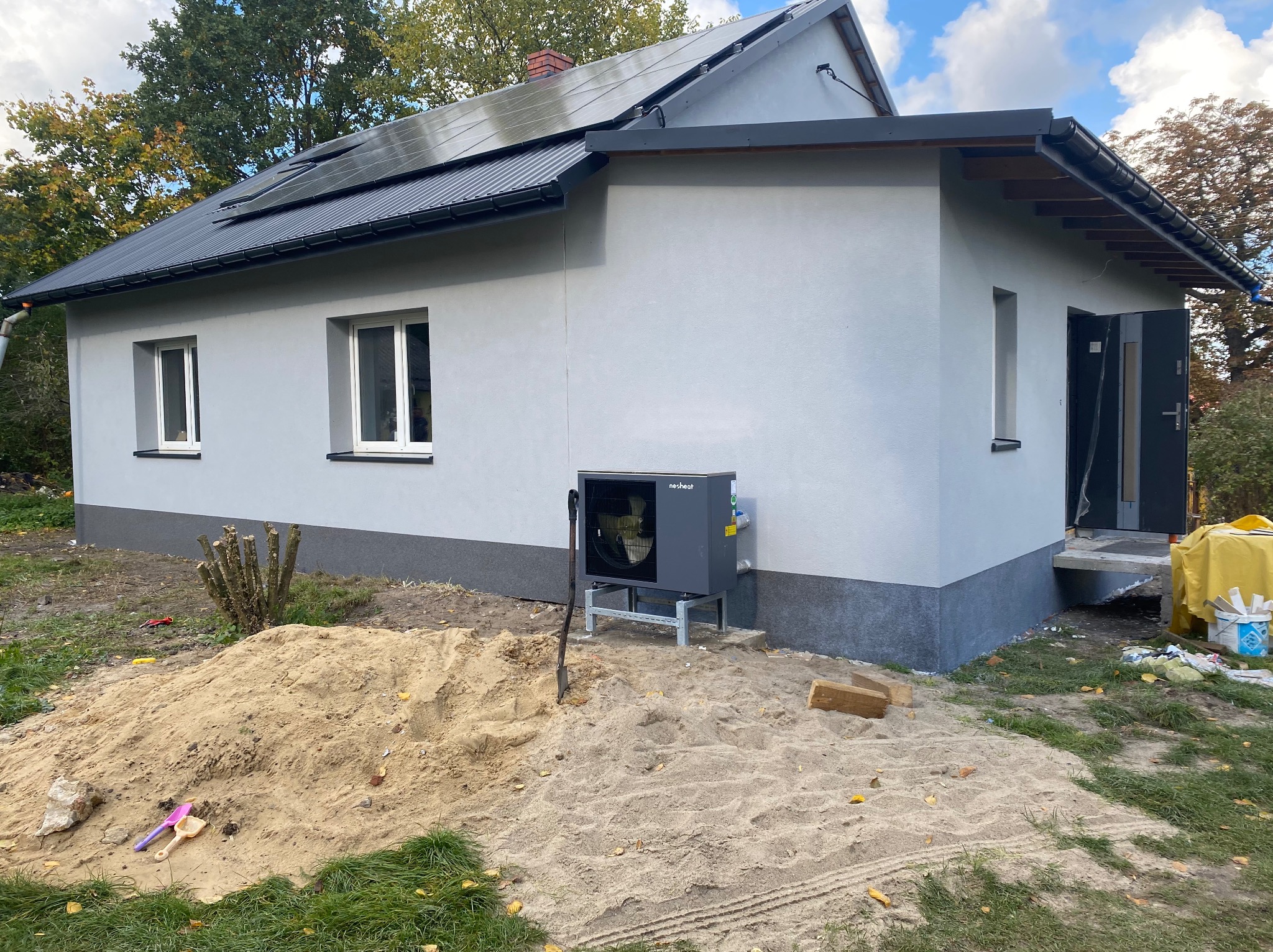 Nowoczesny dom z szarą elewacją, panelami słonecznymi na dachu i pompą ciepła na zewnątrz, widoczny stos piasku i narzędzia, sugerujące prace wykończeniowe.