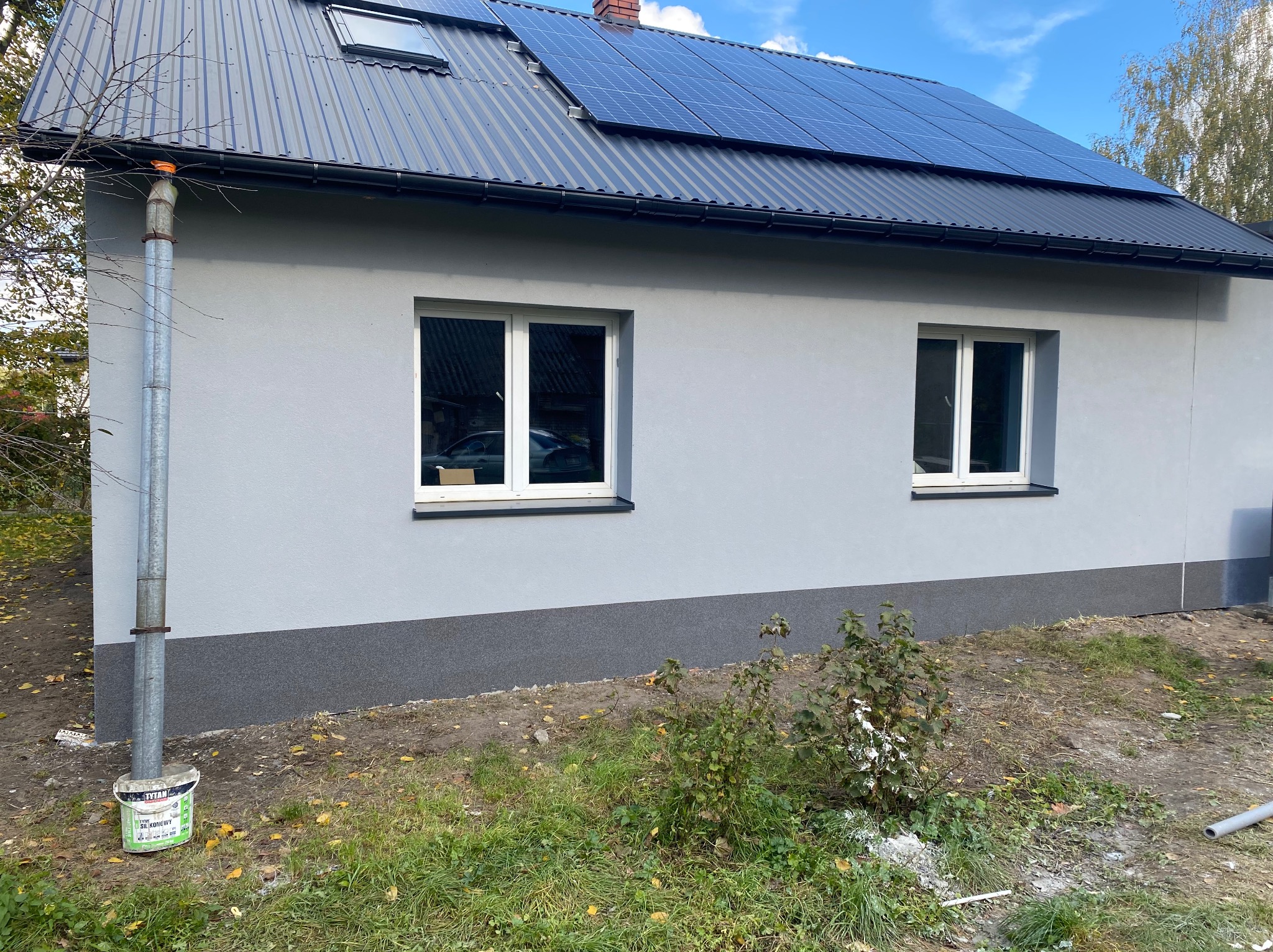 Świeżo otynkowana szara elewacja domu jednorodzinnego z dwoma białymi oknami, ciemnoszarą blachą na dachu z panelami słonecznymi i orynnowaniem, widoczny fragment posesji z roślinnością i wiadro...