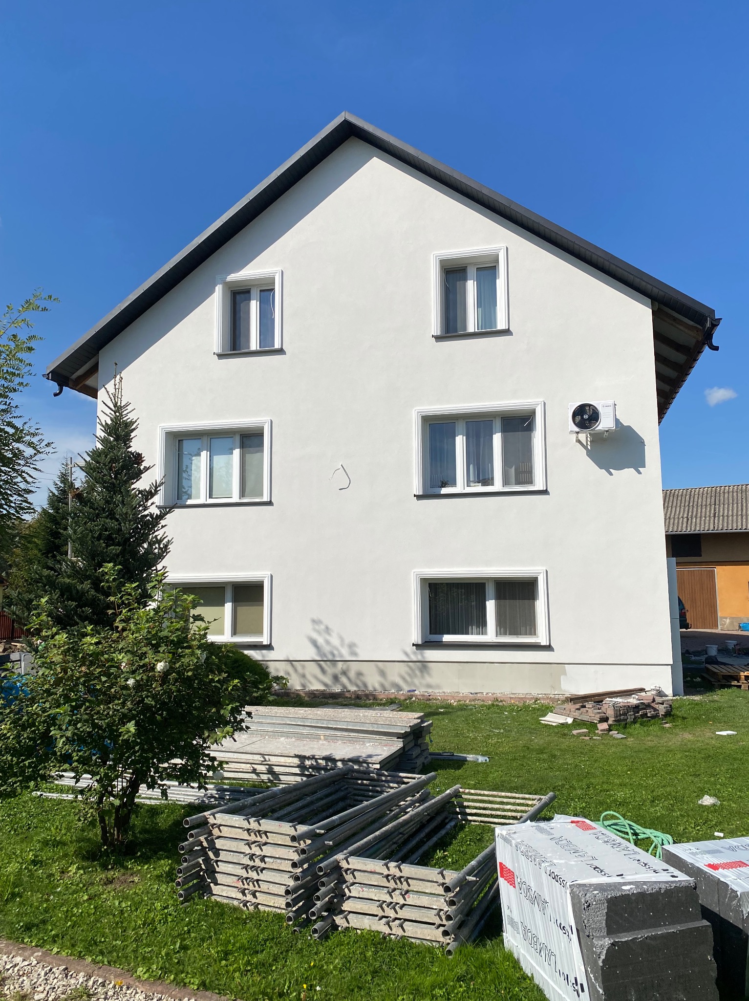 Dwupiętrowy dom z białą elewacją, widoczne okna z białymi ramami, klimatyzator na ścianie, na trawniku leżące rusztowania i paczki styropianu, błękitne niebo.