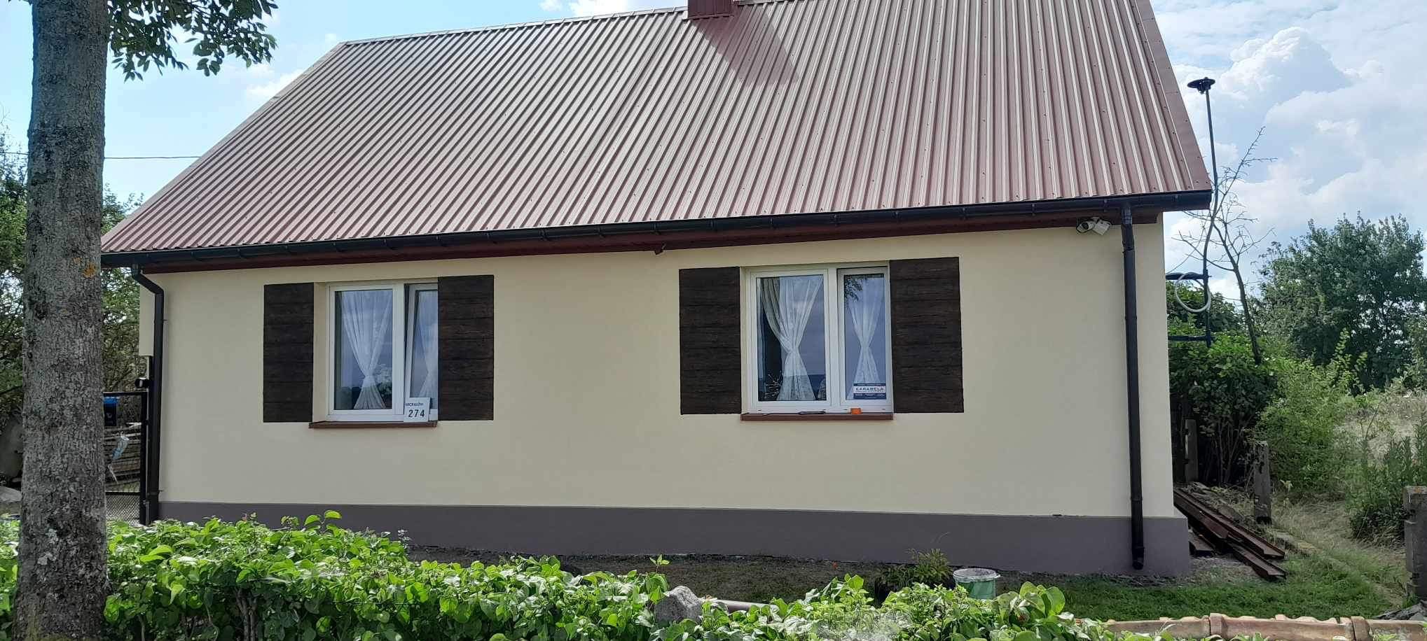 Dom w Tuczępach po renowacji elewacji, z brązowymi okiennicami i beżową ścianą, widok z przodu, na oknach wiszą białe firanki.