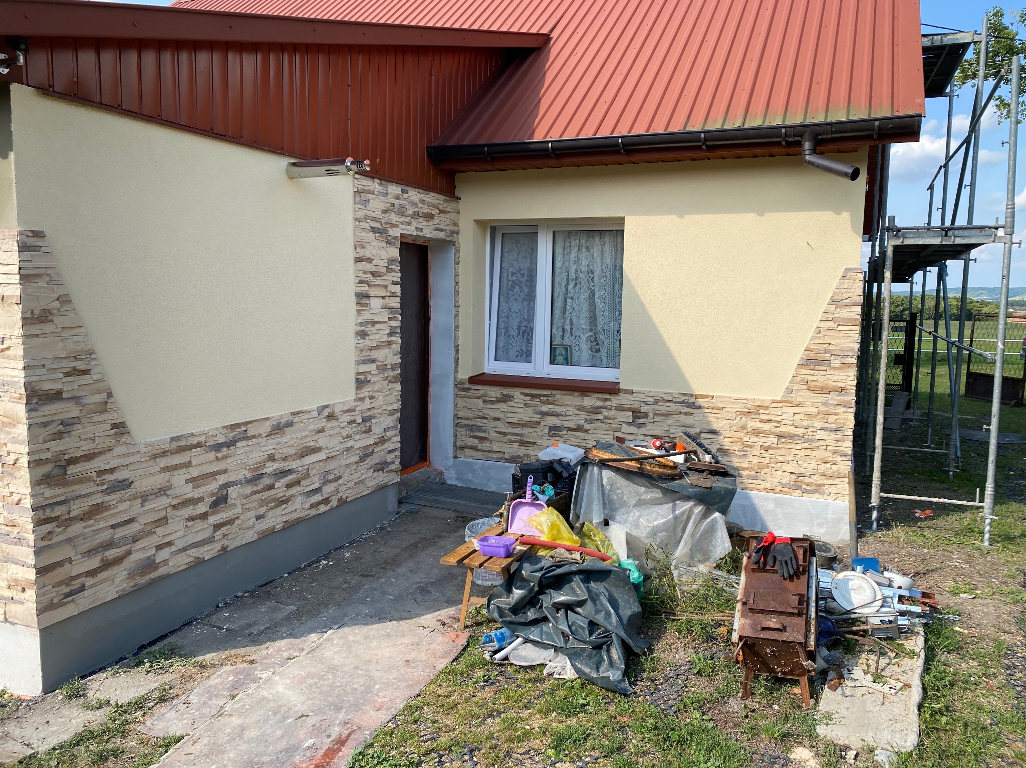 Fragment elewacji domu z ociepleniem wykończonym imitacją kamienia, widoczne rusztowanie, narzędzia i materiały budowlane w nieładzie na trawniku.