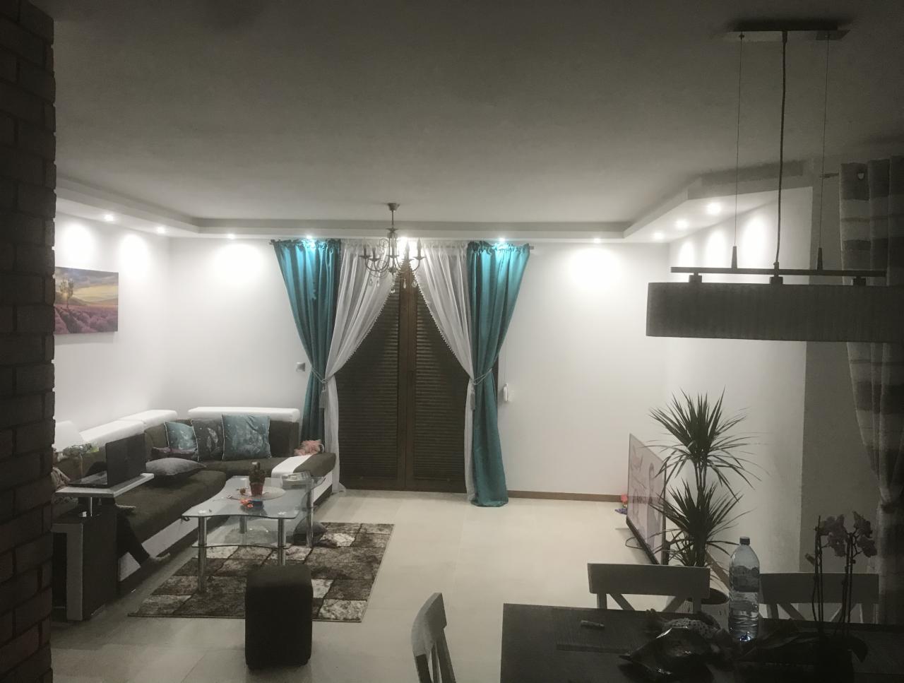 Jasne, przestronne wnętrze salonu z narożną sofą, szklanym stolikiem kawowym, turkusowymi zasłonami i nowoczesnym oświetleniem sufitowym. Na ścianie widoczny obraz z motywem lawendy. Uwagę zwraca...