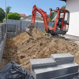 Czerwona mini koparka Kubota na placu budowy, przygotowująca teren pod fundamenty, obok ułożone bloczki betonowe, widoczny fragment ogrodzenia i elewacji budynku w słoneczny dzień.