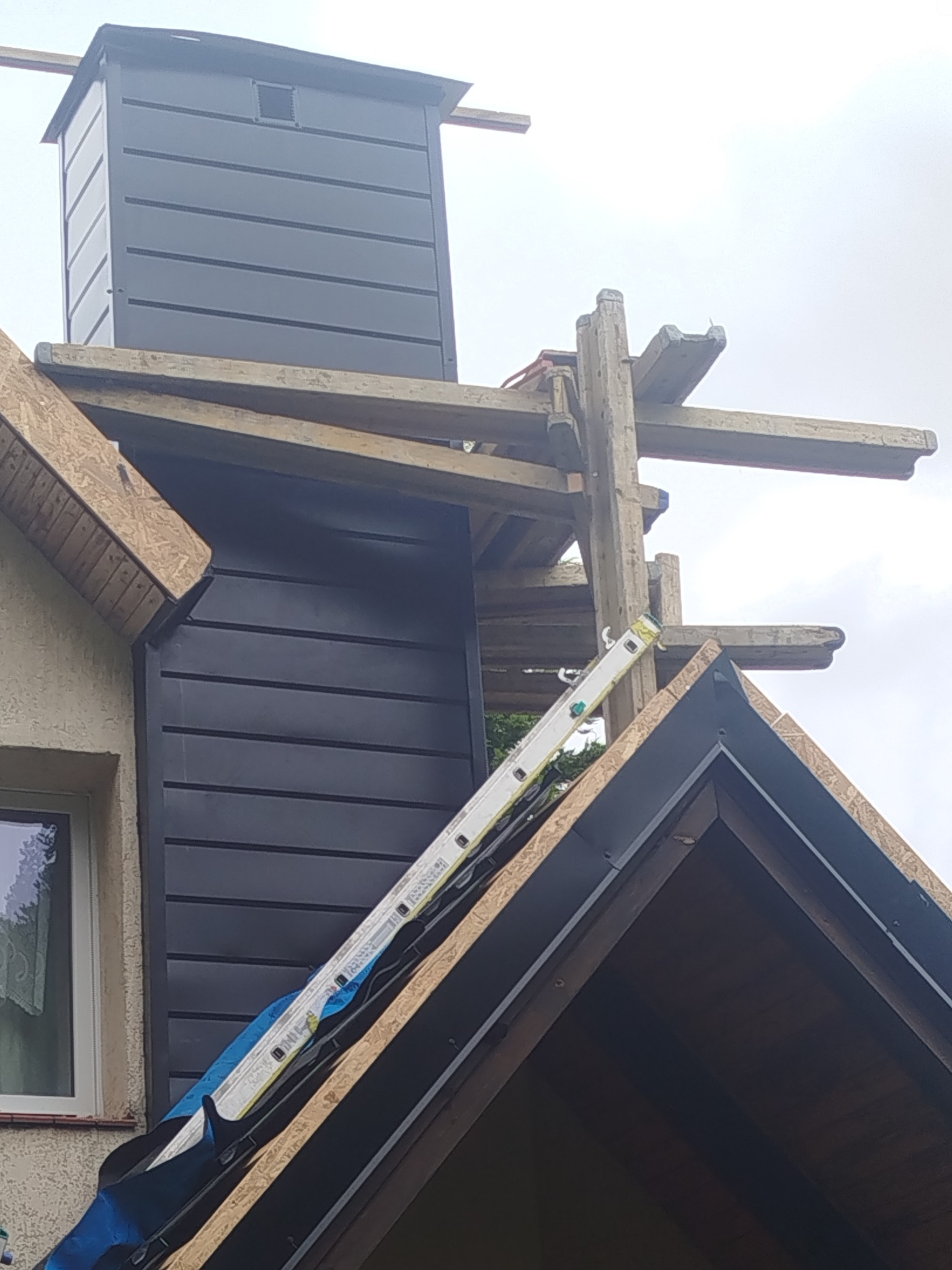 Fragment dachu z kominem w trakcie obróbek blacharskich, widoczna drabina oparta o dach i drewniane rusztowanie. Ujęcie z dołu, pochmurne niebo w tle.