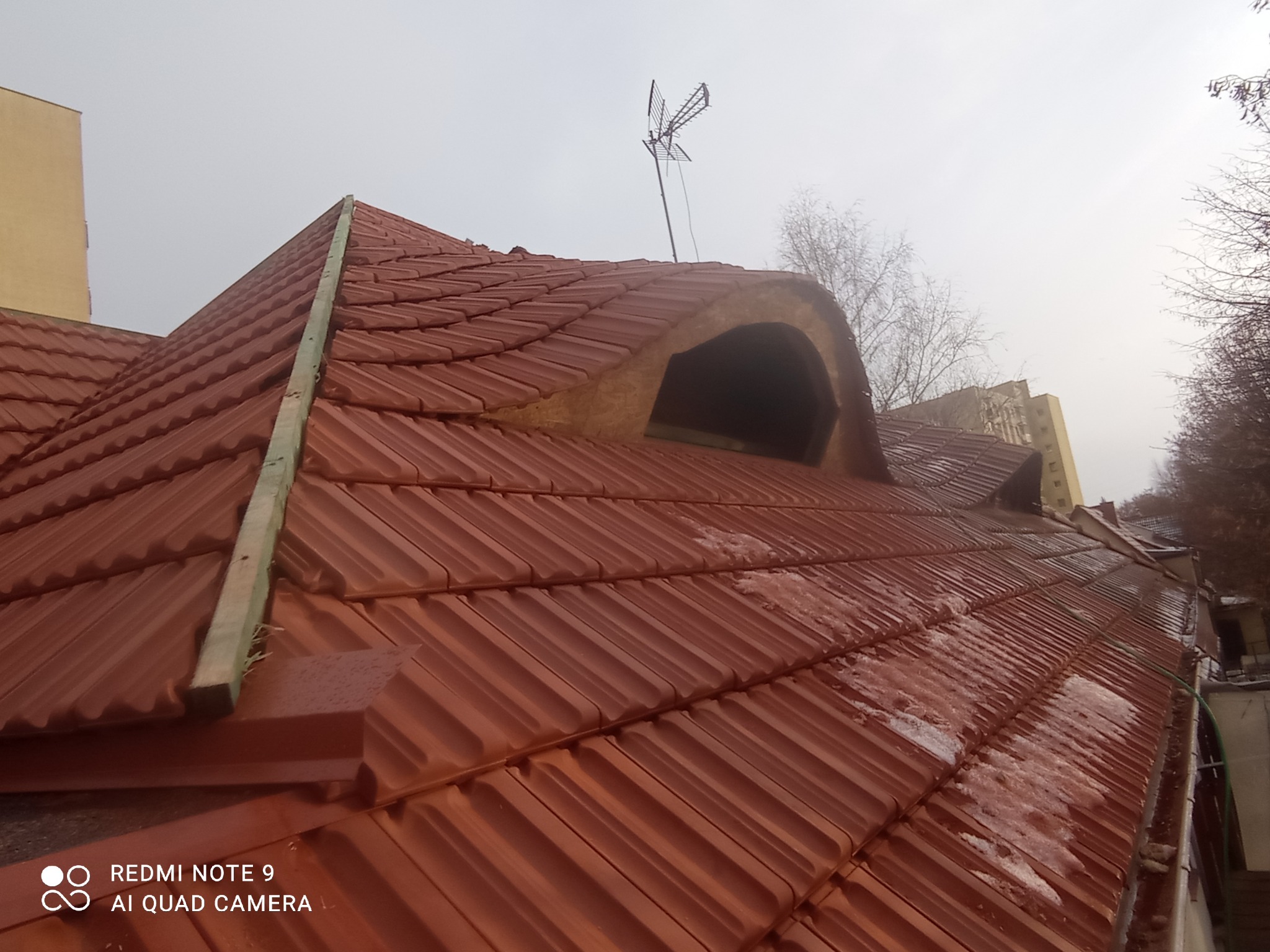 Czerwona dachówka karpiówka na skośnym dachu z lukarną i anteną TV. Widoczne ślady wilgoci i drewniana łata. Pochmurne niebo w tle.