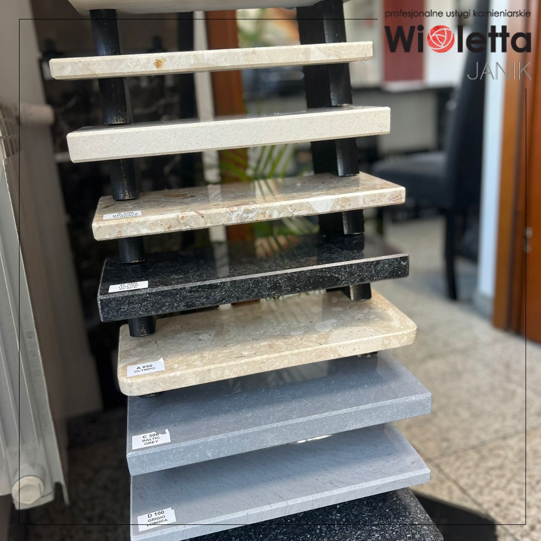 Ekspozycja próbek kamieni naturalnych: granity, marmury w odcieniach bieli, beżu, szarości i czerni. Widoczne etykiety z nazwami kamieni. Aranżacja wewnątrz pomieszczenia.