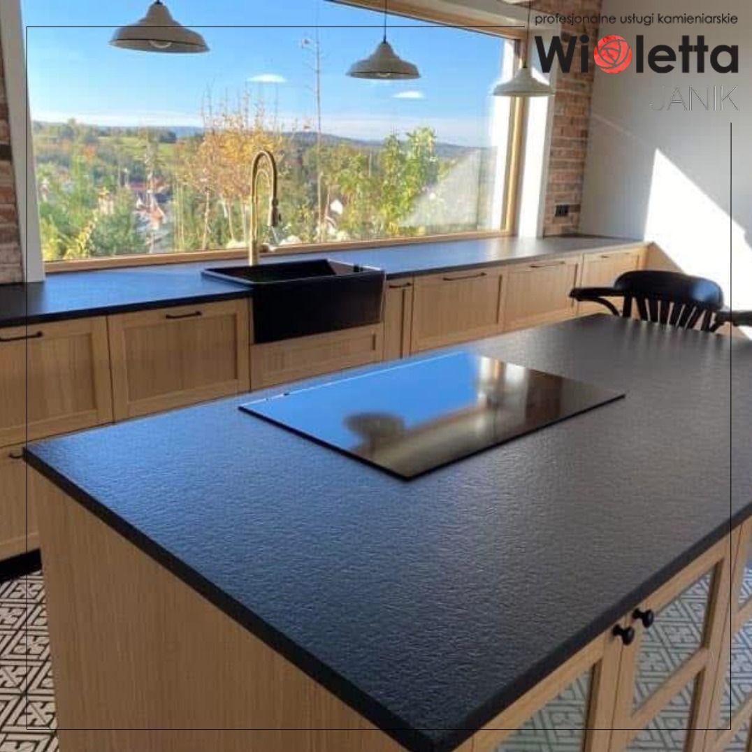 Elegancka kuchnia z blatem z ciemnego granitu, czarnym zlewem farmerskim i widokiem na pagórkowaty krajobraz przez szerokie okno.